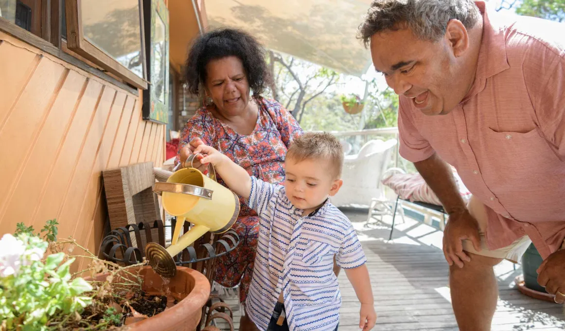 A young boy waters a pot plant while a mature woman and man look on. All stand on a porch with outdoor furniture and trees in the background.