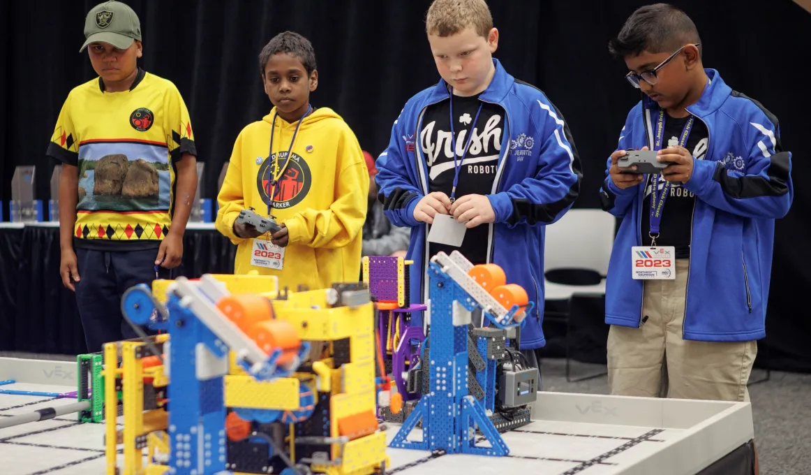 Two Indigenous primary school kids in yellow uniforms compete against two kids from an Irish team, dressed in blue. Their robots are in the foreground of the photo.