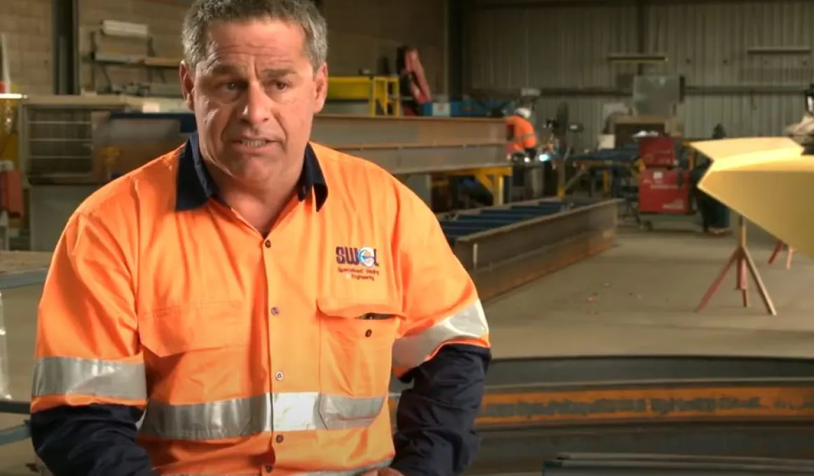 A middle aged man in orange and blue work wear sits in a large workshop. In the background is heavy machinery, a worker and fabricated metal in various shapes.