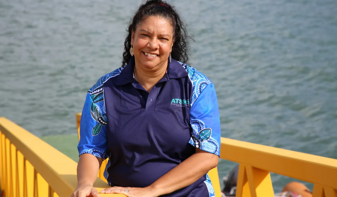 Aboriginal woman in blue shirt stands next to a yellow rail with the ocean in the background.