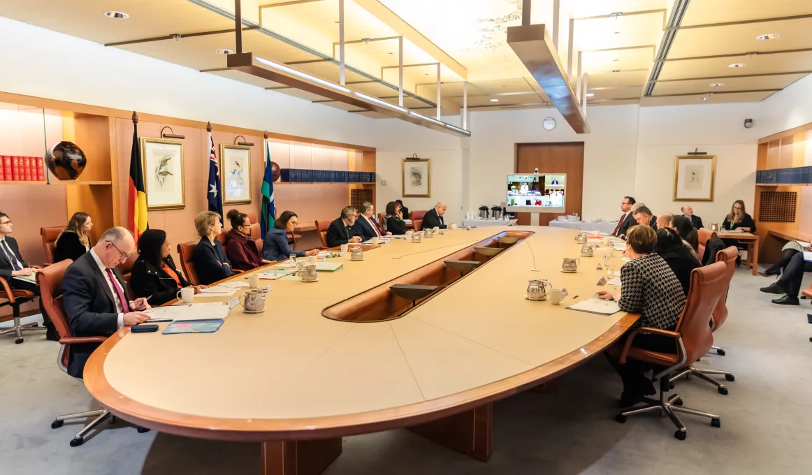 Group of people formally dressed sit around a board table in a large room. In the background is wood pannelling, more people, a door, flags and other office content.