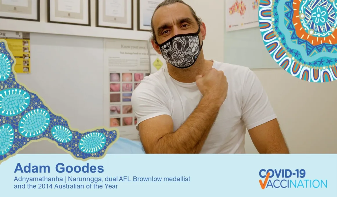 A man sits in a Doctors office. He is wearing a white t-shirt and a black and white patterned face mask. He has one arm held across his chest to show where his upper arm where he was vaccinated