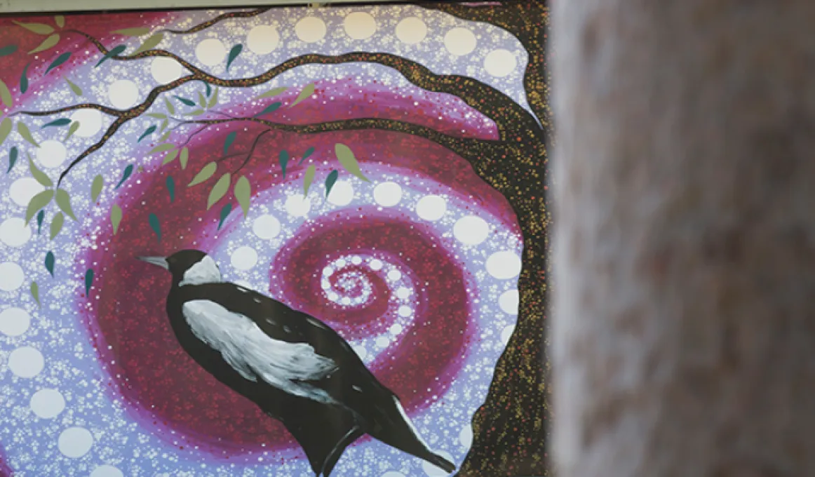 A black and white bird stands next to the trunk of a tree. In the background is a white and moroon swirl made up of circles and dots. In the foreground is another tree trunk.