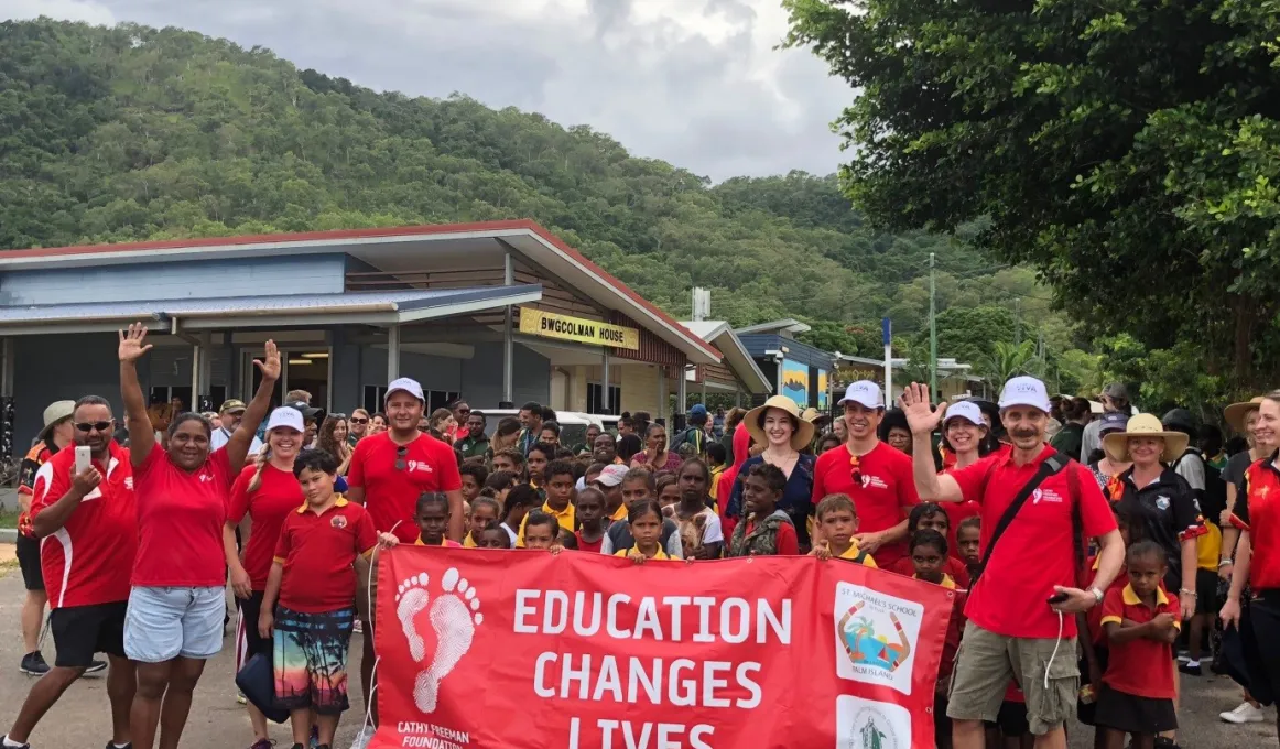 A group of men, women and children in red school polo shirt with a crowd of Indigenous children behind red banner with white writing on banner ‘Education Changes Lives.’