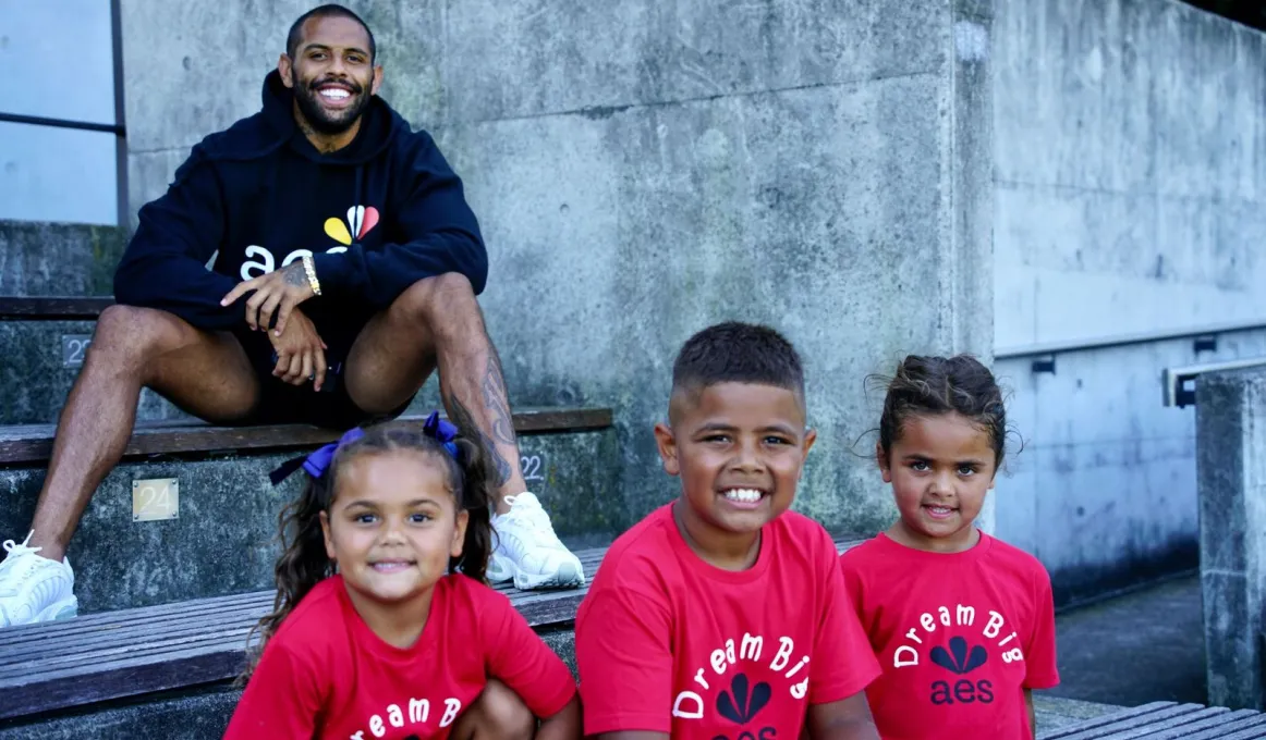 A man in black top and shorts sits on a step in an outdoor area. Three children in red tops sit below him.