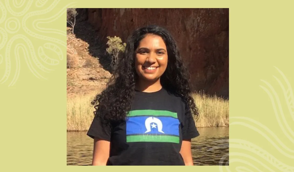 Indigenous woman with long black hair wearing a black t-shirt with a blue, green and white flag on the front stands in front of body of water. In the background is grass and a cliff face.