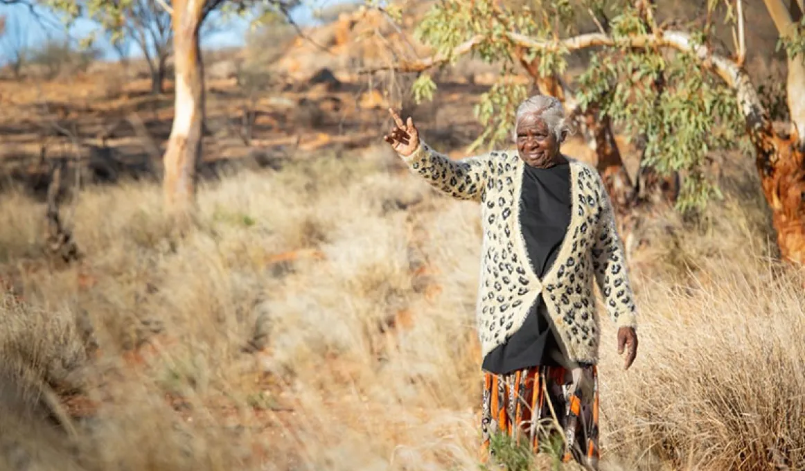 A woman wearing a black top, orange floral skirt and leopard print cardigan is walking in the scrub surrounded by trees. The woman is indicating to something in the distance that is not visible in the image.