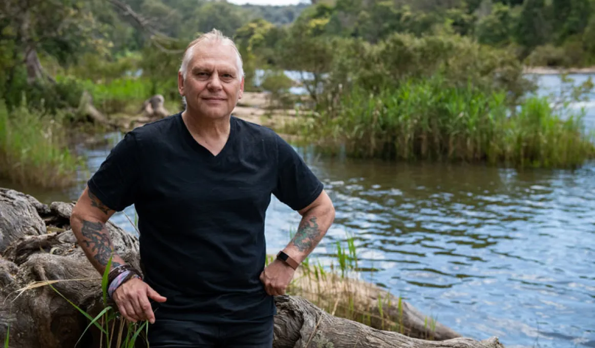 A man wearing a black shirt and blue jeans is standing on the edge of a river. Behind him are some rocks and trees.