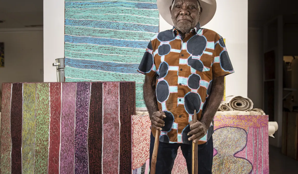 A man wearing a blue, pink, brown and white patterned shirt is standing in front of a collection of brightly coloured artwork. He is wearing a hat and is holding two, long carved sticks.