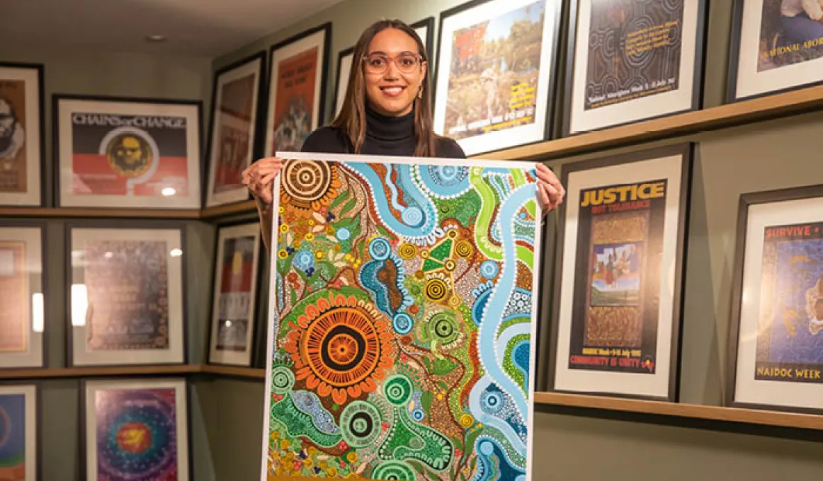 National NAIDOC Week winning artist Maggie-Jean Douglas holds a copy of her winning NAIDOC Week poster. In the background are framed versions of previous NAIDOC Week posters on the wall.