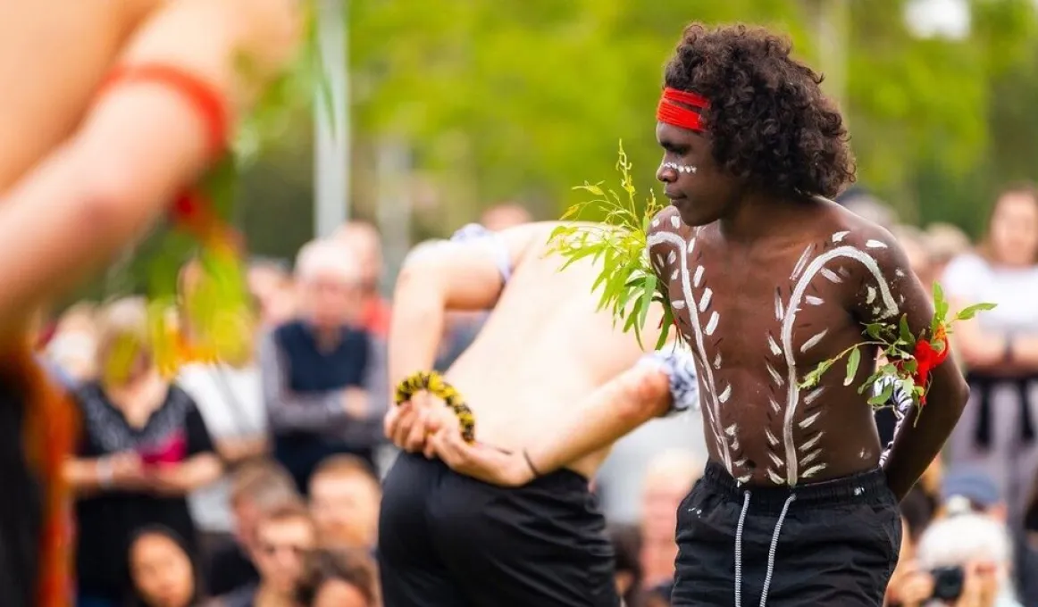 Three men dancing in front of a crowd. The dancers are wearing black shorts, white paint on their chests and coloured armbands. Each arm band is holding a small branch with green leaves.