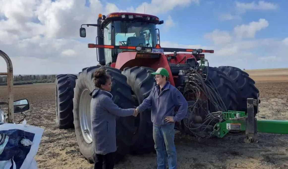 A woman in a long grey coat shakes hands with a man in jeans and top. In the background is a tractor and open field.