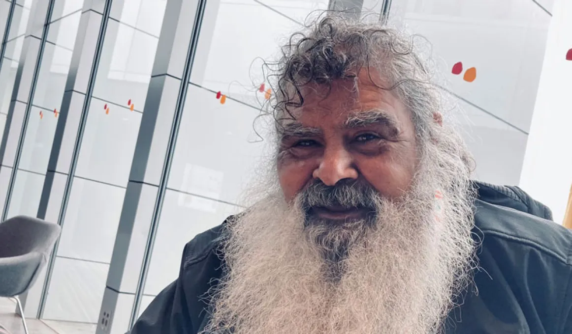 An older man with a long, white beard, a curly fringe and a black jacket sits in front of a glass wall.