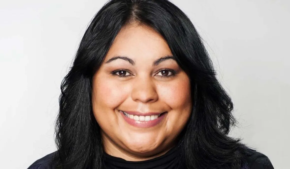 A woman with black hair and brown eyes smiles at the camera. She is wearing a black turtle neck top with black lace detailing.