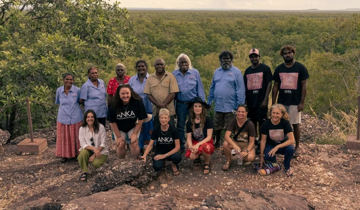 A group of people dressed in casual or work wear stand or squat on stony soil near the edge of a drop off. In the background is a tree covered plain and cloudy sky.