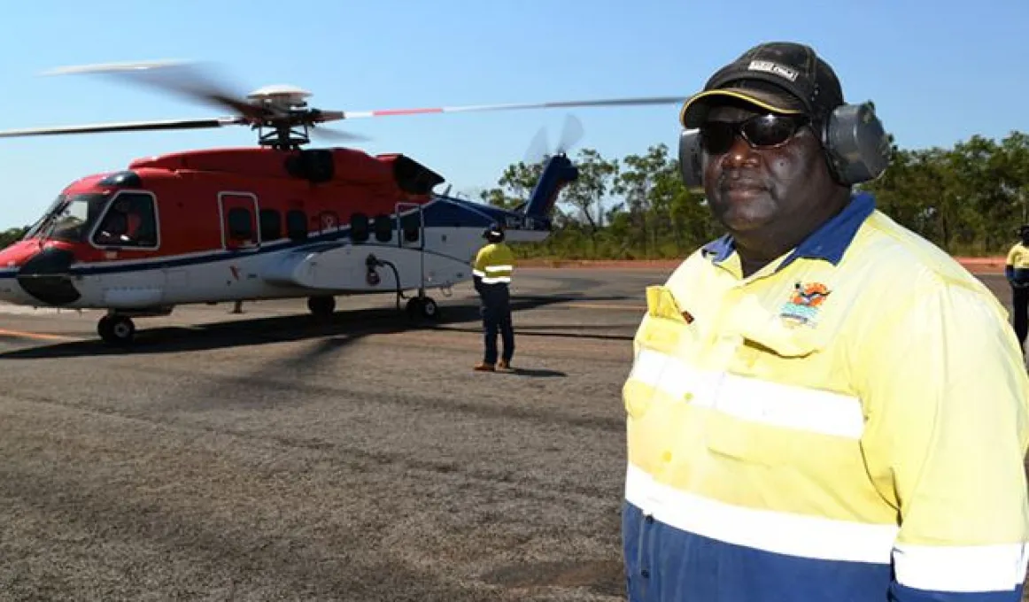 A man in work wear and including safety headphones and a cap stands on an aircraft tarmac. In the background are other men and a large helicopter. Behind that is bush land.