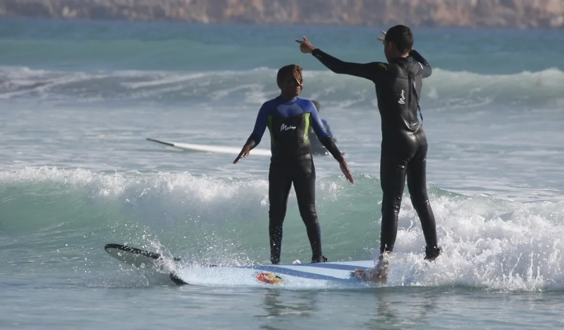 Two Aboriginal youth in wet suits surf side by side. In the background are waves on an ocean and behind that, a cliff face.