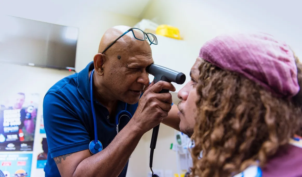 Male doctor in blue shirt uses an instrument to check the eye of another man with dreadlocks and wearing a pink beret. In the background are posters.