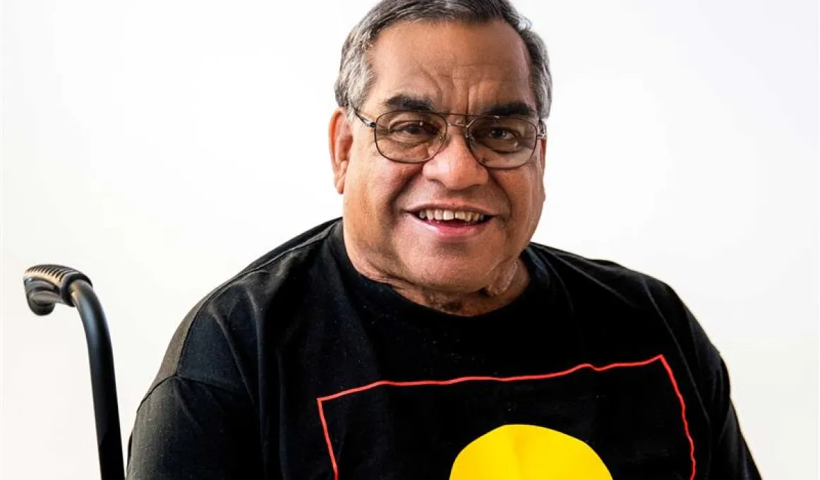 Close up image of a middle aged man sitting in a wheel chair. The man is smiling and wearing glasses and a t-shirt with the Aboriginal flag on it.