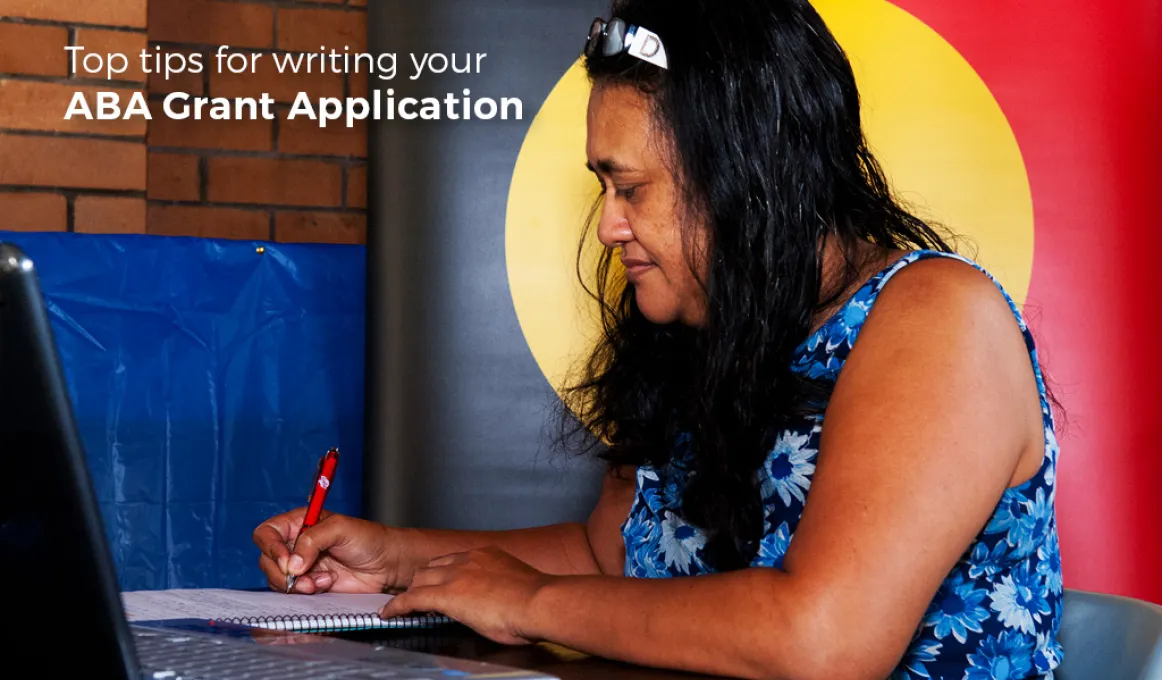 Indigenous woman writing on paper with a pen and the Aboriginal flag in the background, a laptop computer in the foreground and the following words displayed: Top tips for writing your Aboriginal Benefit Grant Application