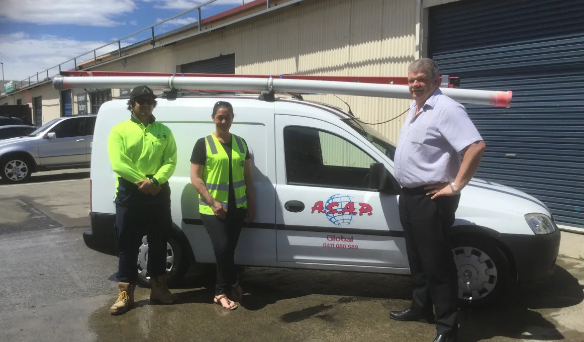 An Indigenous young man and young woman wearing safety tops and an older man dressed in light shirt and dark trousers stand in front of small white van parked in front of yellow building with dark green roller door. Letters on the car door are A.S.A.P.