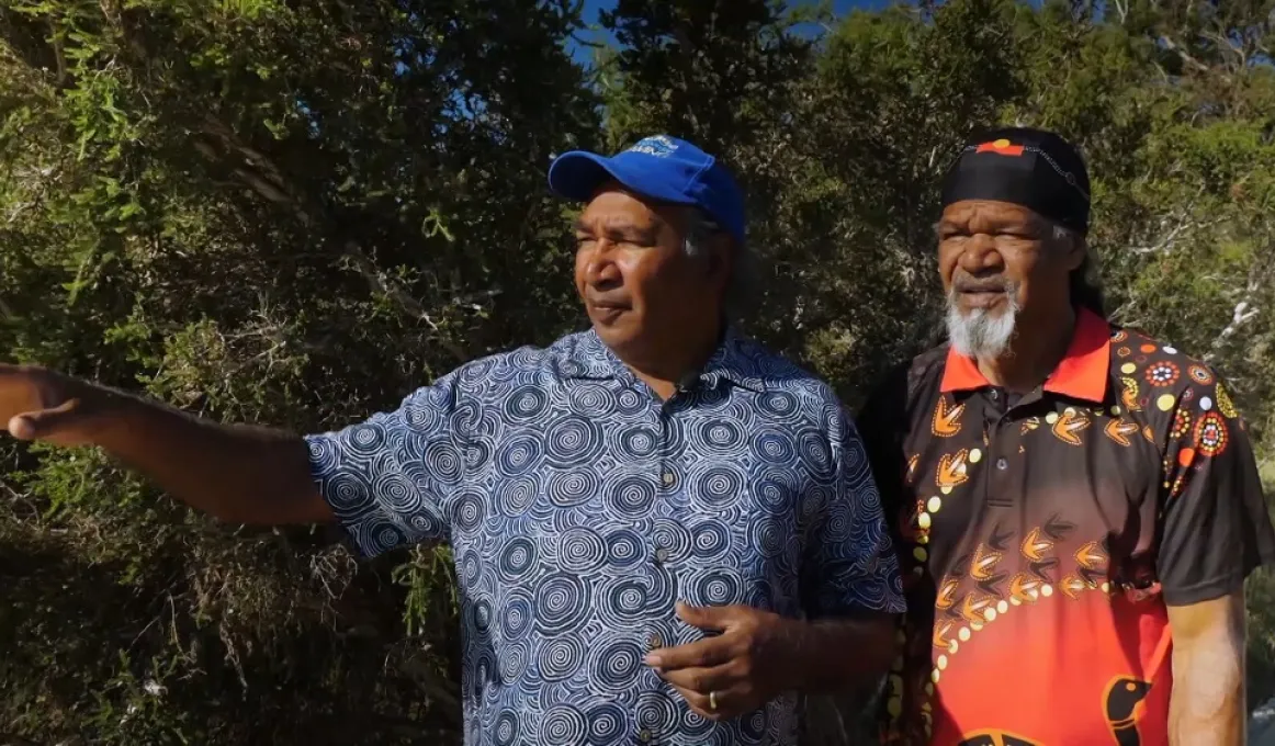 Two Aboriginal men stand side by side. The man on the left points to his right. He wears a blue and white shirt and blue head covering. The man on the right wears a red, brown and yellow shirt and similar coloured head covering.