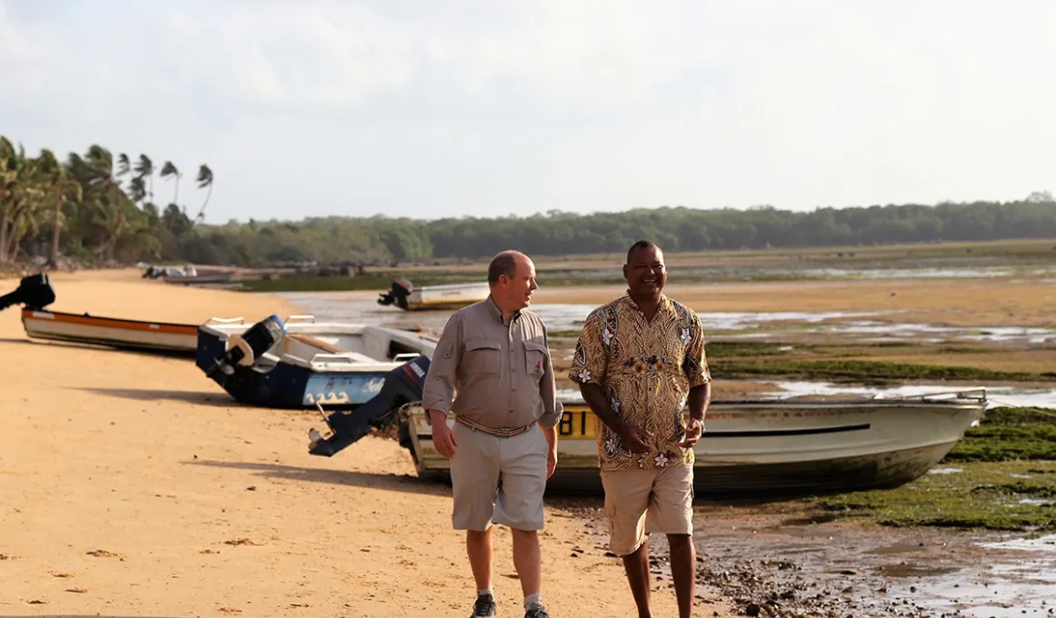 Two men walk along a beach with boats, reef and weed and trees in the background. Both wear shorts and casual shirts.