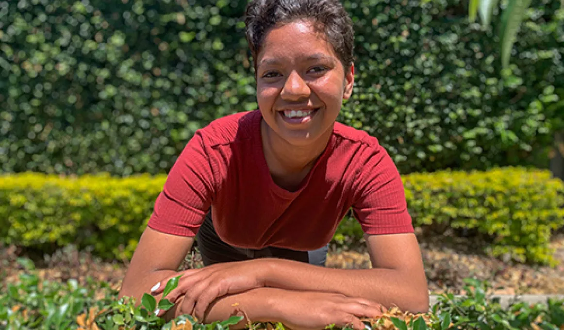 Young woman with short hair and red shirt leans on a hedge and faces the camera. In the background are more hedges.