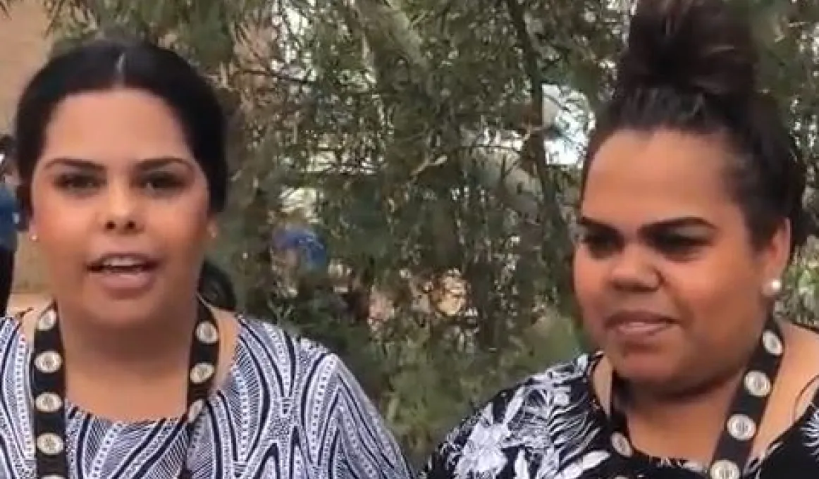 Two Indigenous women in black and white clothing stand in front of trees.