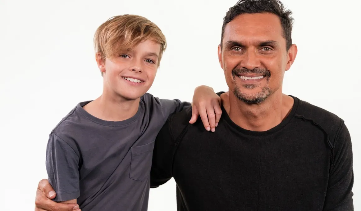 A boy with light coloured hair and grey t-shirt has placed his hand on the shoulder of a man with black hair and a black t-shirt. The man has placed his arm around the boy.