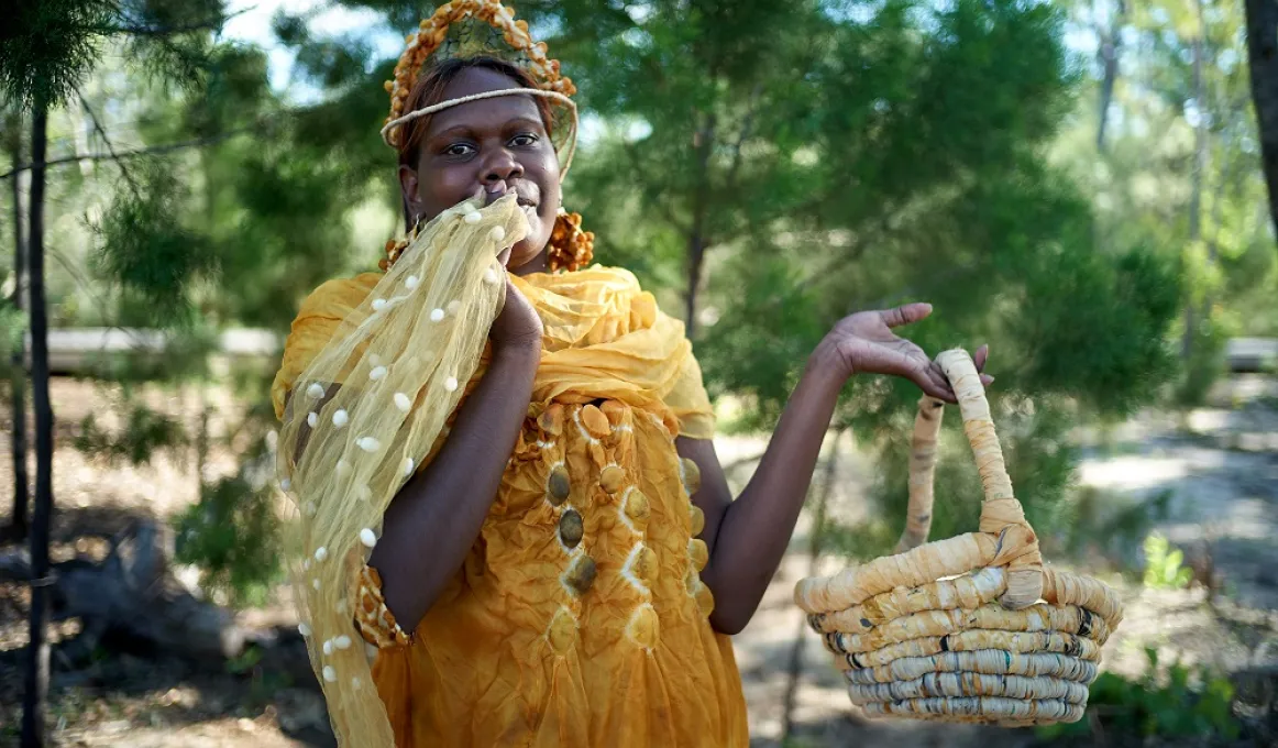 An Indigenous woman models a golden yellow silk dress, scarf and headpiece, holding up in one hand a handwoven-coiled basket.  She is standing in a natural environment, shaded by green trees.