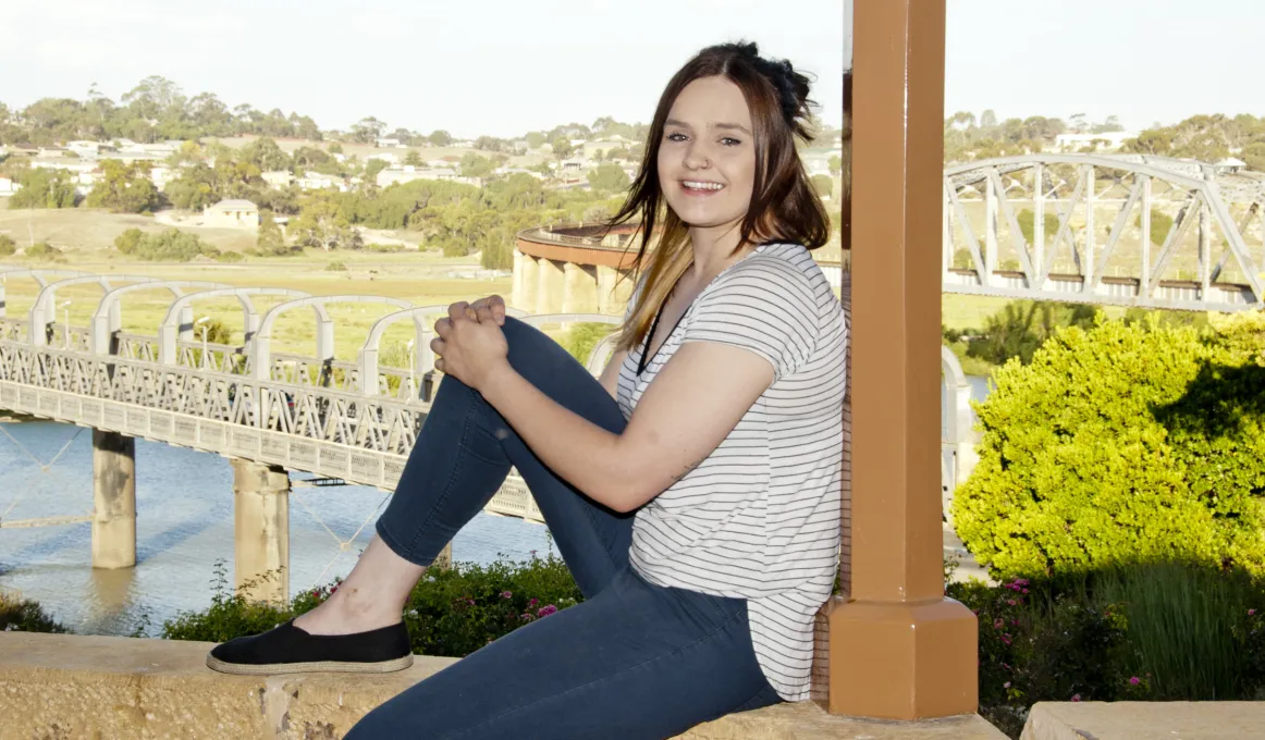 Young woman sits on a wall with her back to a pillar. She is dressed in dark slacks and light shirt. In the background is a tree, a river, two bridges and houses.