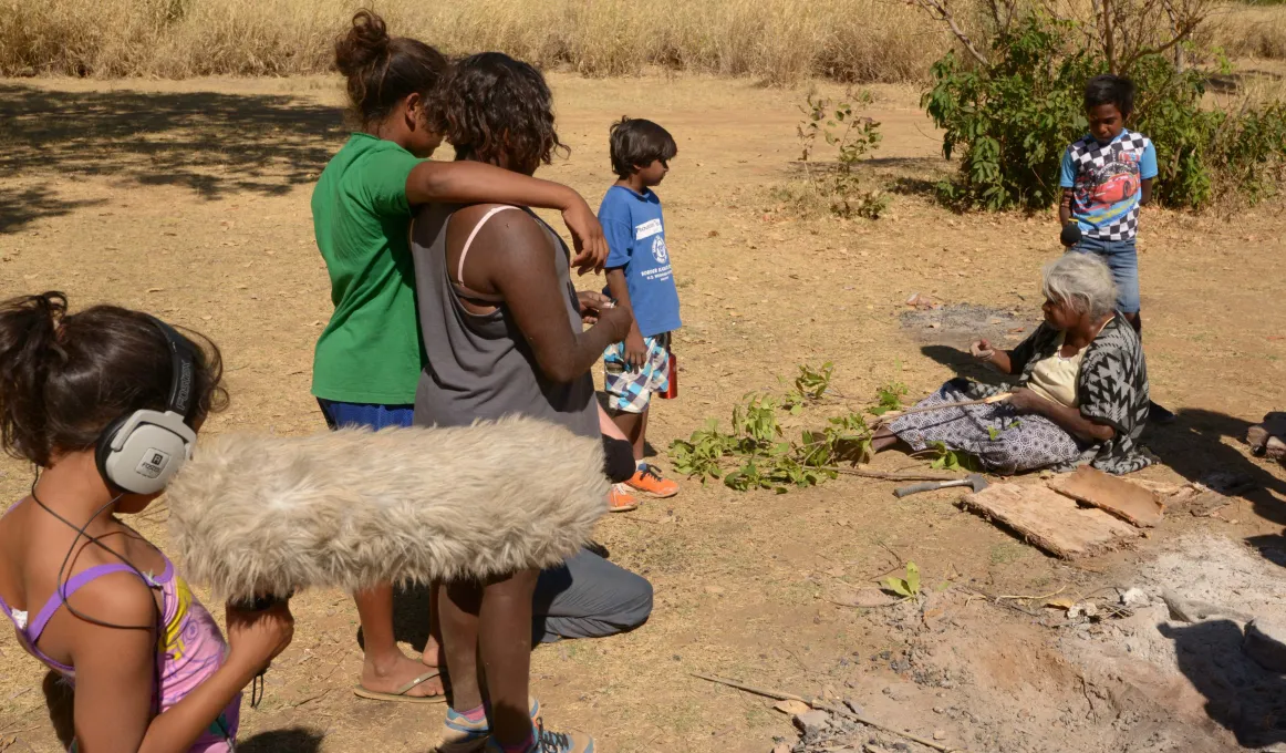 An elderley woman sits on th ground holding a branch. Next to her is bark and a cold campfire. Standing around her a young women and young men. In the background are bushes and long grass and trees.