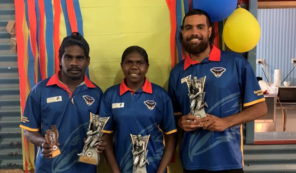 Two young adult men and one woman hold trophies and wear blue polo shirts. In the background is a corrugated wall and red, blue and yellow decorations.