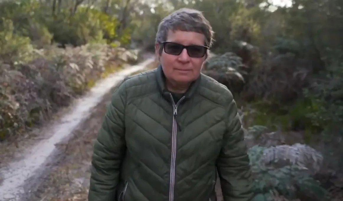 A woman with short hair and wearing sunglasses and a green coat walks along a path. Behind her is the path with shrubs and trees on both sides.