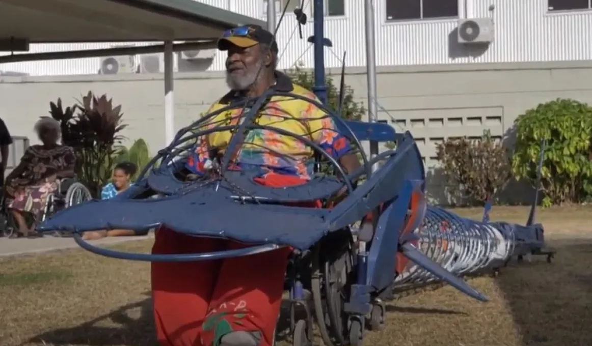 An elderly man sits in a wheel chair that forms part of a long metal contraption in the shape of a hammerhead shark. In the background are two women, grass, shrubbery and a building.