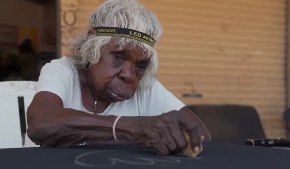 An elderly Aboriginal woman wearing a headband around her grey hair sits at a table and sketches on a black canvas.