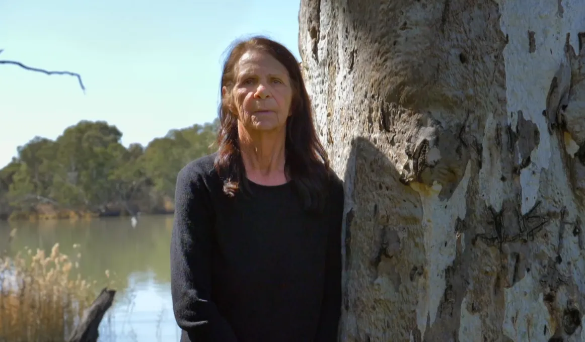 An elderly woman with dark long hair and wearing a dark top stands next to a gum tree. In the background are water and trees.