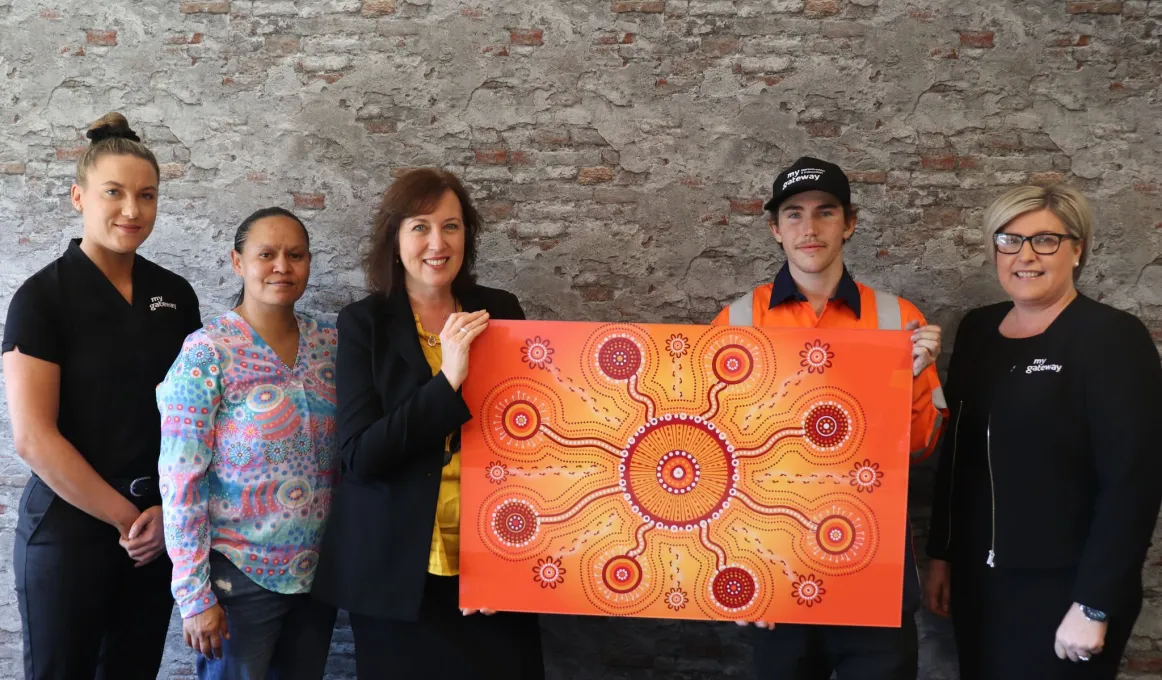 Five people stand in front of a mottled wall. A woman and a young man hold a painting between them. It is predominantly orange and yellow in colour with a circle at centre connected to other circles radiating from the first.