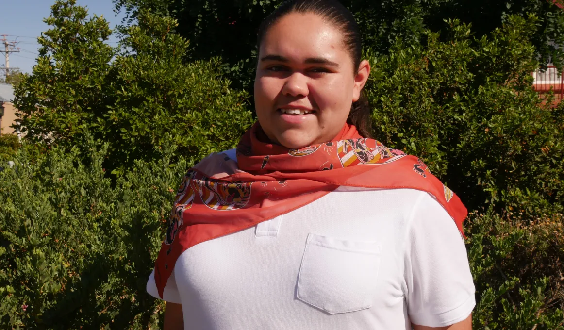 Young woman in white top and ochre coloured scarf stands in front of trees and bushes.