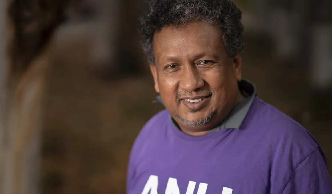 Aboriginal man with curly dark hair wearing a purple top with the letters A.N.U. on the front.