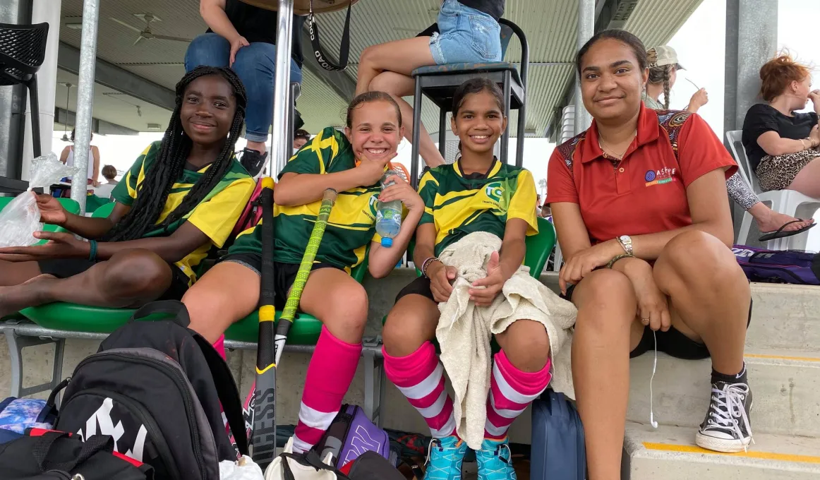 Three young Indigenous women sit in their green and yellow hockey apparel with an older young woman in an ochre coloured shirt. In the background are more people sitting under cover.