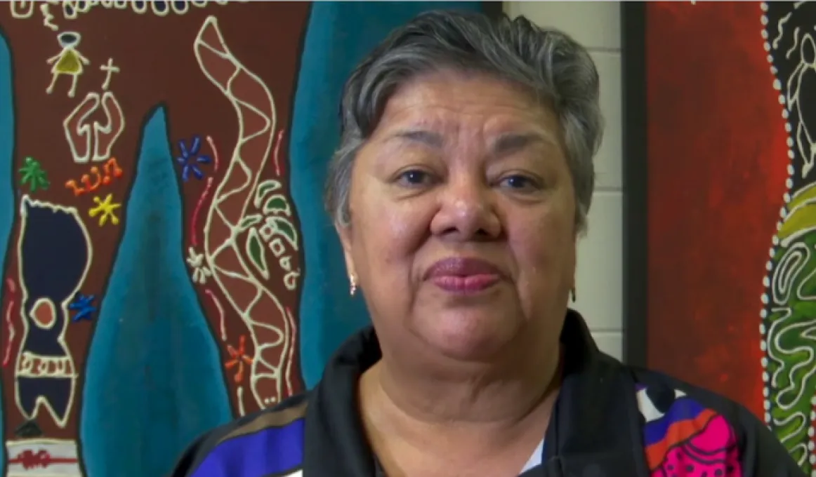 A woman sits in front of two brightly coloured Indigenous artworks. She is wearing a black and brightly coloured polo shirt.