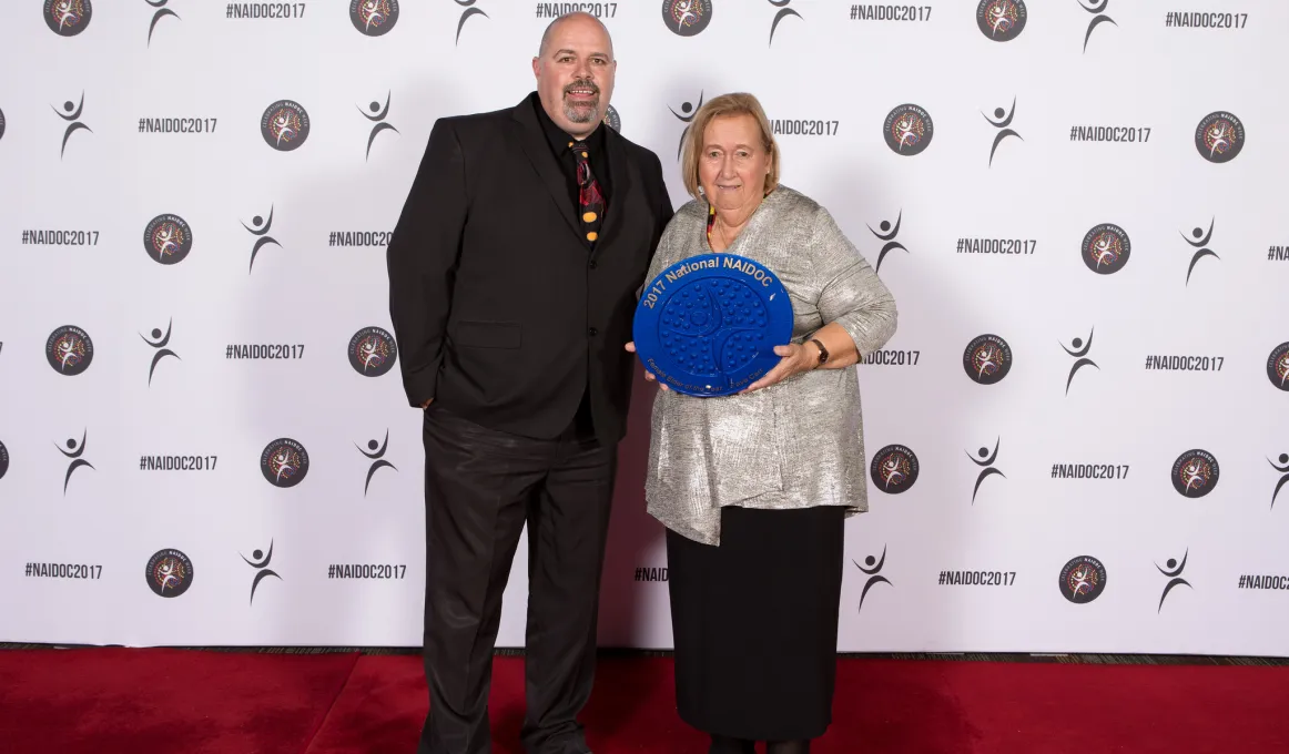 Indigenous male stands next to an older Indigenous woman on red carpet, the woman is holding large blue plate.