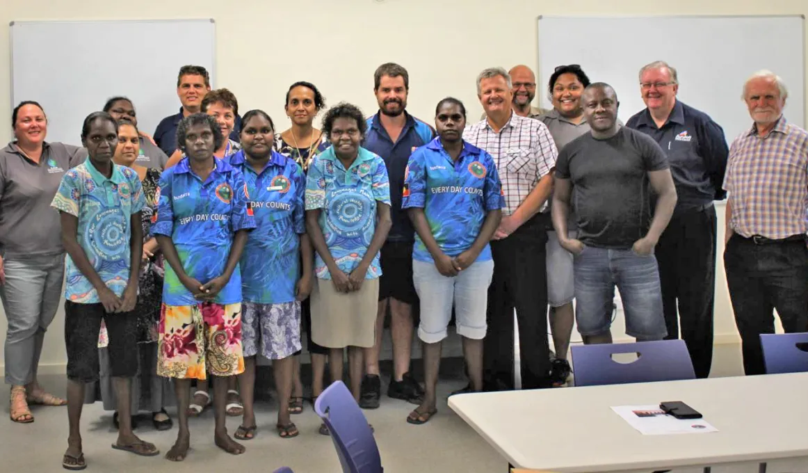 Large group of Indigenous and non-Indigenous men and women dressed in casual clothing in a room with beige carpet. In the background is a pale yellow wall and two whiteboards. In the foreground is a white table and blue chairs.