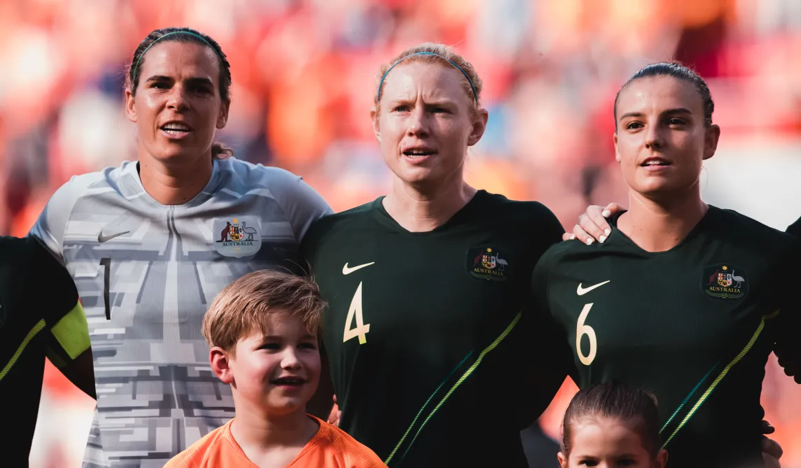 Three women (one in grey shirt and two in dark green shirts) stand arm in arm with two children in front of them. In the background is a crowd of people mainly in orange clothing.