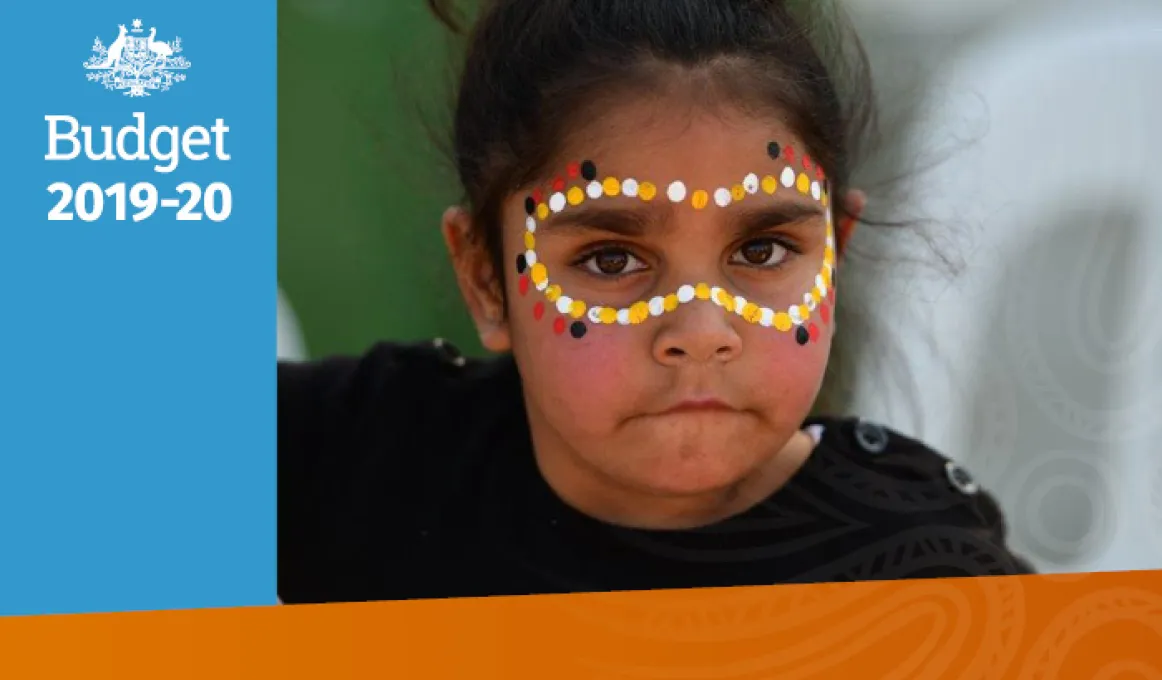 A primary-school aged girl looks at the camera. She is wearing a black and white striped jumper. She is wearing yellow, white, red and black face paint in the traditional dot style around her eyes.