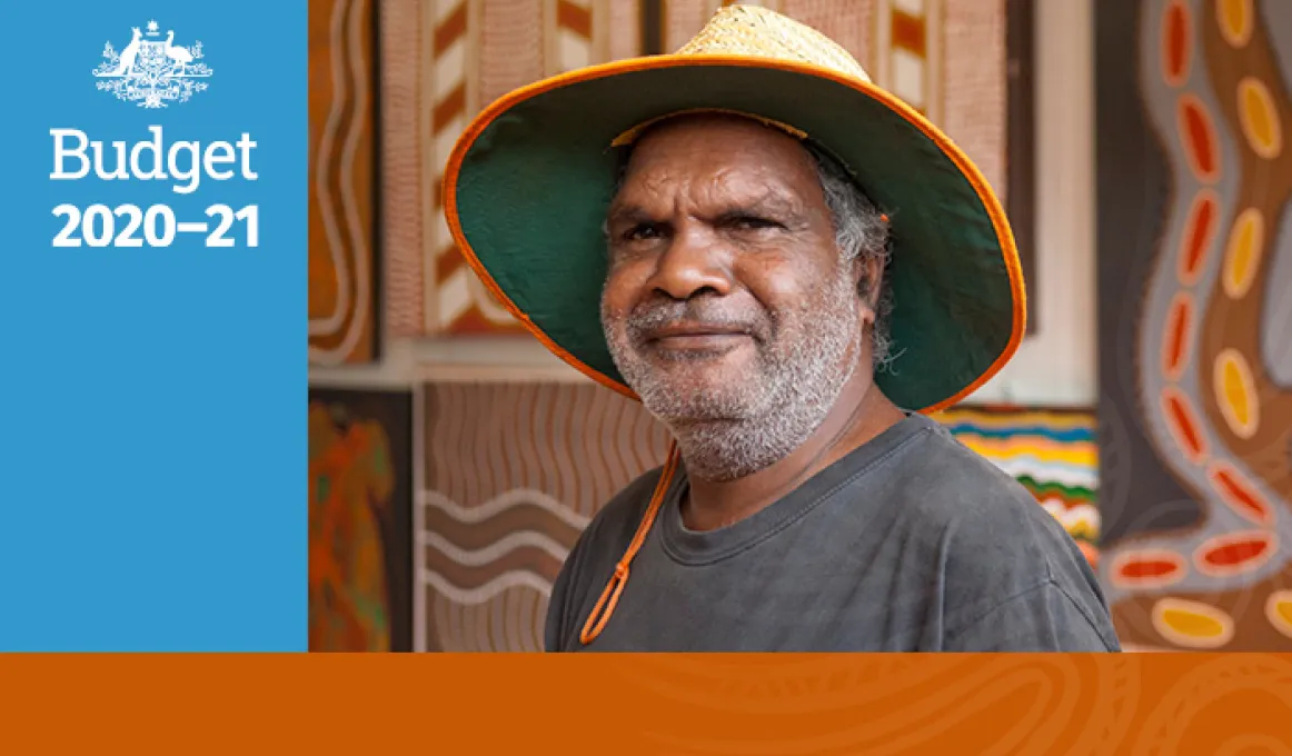 A man wearing a grey shirt and a wide brimmed straw hat is standing in front of Indigenous artwork comprised of wavy and straight lines and red and yellow large dots. Beside there is a blue rectangle with Budget 2020 – 2021 and the Australian Government C