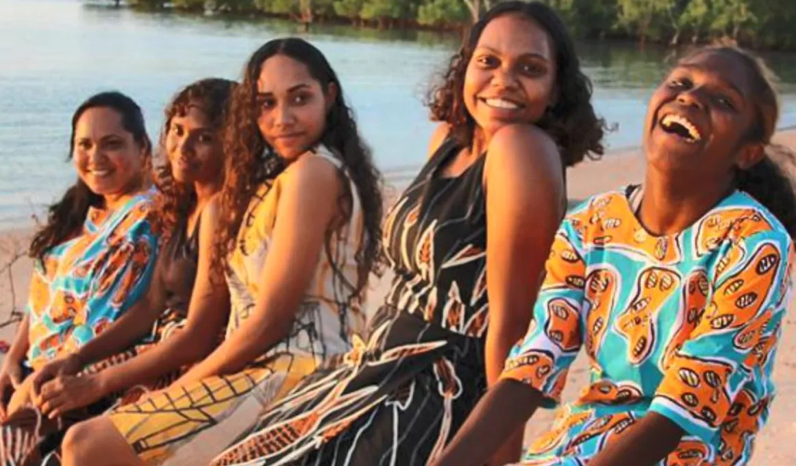 Five Indigenous women wearing colourful clothing sit in a line on a beach with water and trees in the background.