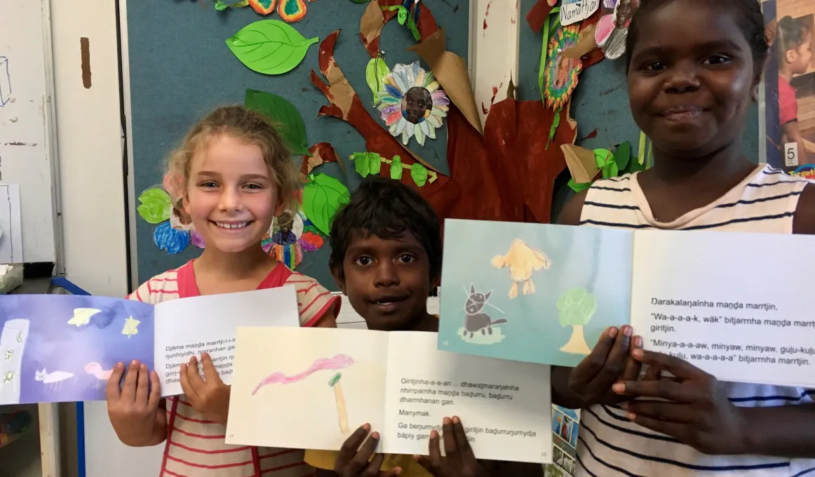 Three children stand in front of a colourful artistic display. Each holds a children’s book open to a two page spread with paintings on the left and text on the right.